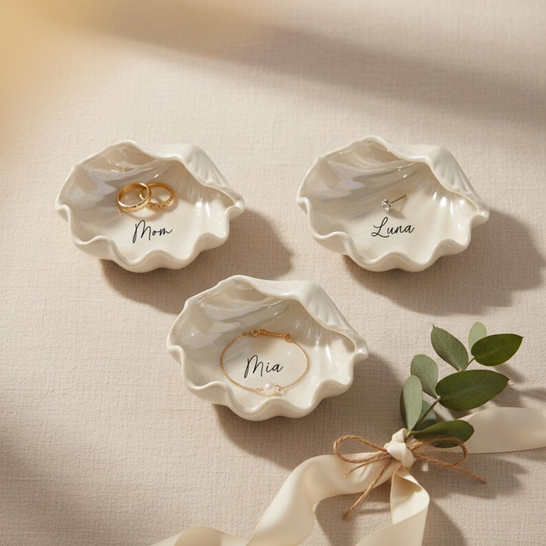 May include: Three white ceramic seashell-shaped ring dishes. One dish holds two gold rings and the word "Mom". Another holds a single earring and the name "Luna". The third holds a gold bracelet and the name "Mia".