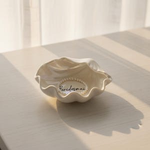 May include: A white, seashell-shaped trinket dish with a pearl bracelet inside. The dish has a wavy, textured rim and the word "Bridesmaid" is written in script. The dish is on a light-colored surface.