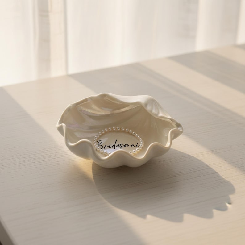 May include: A white, seashell-shaped trinket dish with a pearl bracelet inside. The dish has a wavy, textured rim and the word "Bridesmaid" is written in script. The dish is on a light-colored surface.
