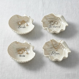 Pode incluir: Quatro pequenas travessas em forma de concha, em branco sujo. Cada travessa tem uma inscrição diferente em escrita preta: "Bride", "Bridesmaid" e "Mom". As travessas contêm joias, incluindo brincos, anéis e um colar.