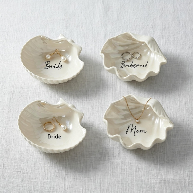 May include: Four off-white seashell-shaped trinket dishes. Each dish has a different inscription in black script: "Bride", "Bridesmaid", and "Mom". The dishes hold jewelry, including earrings, rings, and a necklace.