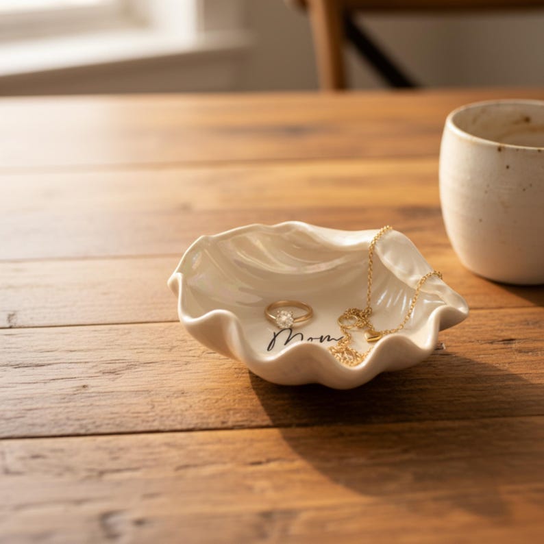 May include: A cream-colored, seashell-shaped ceramic jewelry dish with a gold ring and necklace. The dish has the word "Mom" written in cursive. A white mug sits in the background on a wooden table.