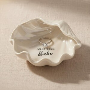 May include: A white ceramic seashell-shaped ring dish with a silver ring. The dish has the date "05.12.2023" and the word "Babe" printed on it. The ring has a large, clear gemstone.