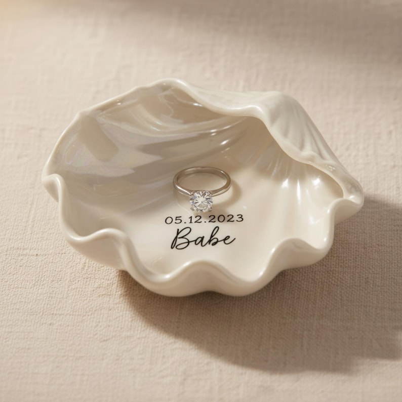 May include: A white ceramic seashell-shaped ring dish with a silver ring. The dish has the date "05.12.2023" and the word "Babe" printed on it. The ring has a large, clear gemstone.