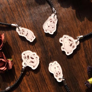 May include: Five clear acrylic charms featuring illustrated cartoon characters with small flower details, each attached to a black braided cord strap with a silver-toned metal connector. The charms display various whimsical character poses.