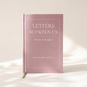 May include: A dusty rose hardcover journal with white text that reads LETTERS BETWEEN US Mother & Daughter written back and forth, kept for years. The book features a thin white rectangular border and a small green ribbon bookmark at the bottom.