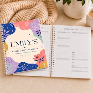 May include: A spiral-bound daily planner with a colourful abstract floral cover featuring the text EMILY'S ADHD DAILY PLANNER a gentle system for busy minds. The open page displays a structured layout with sections for daily focus, time blocks, and meals.