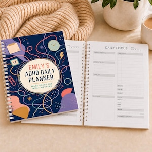 Peut inclure: Un agenda journalier à reliure spirale avec une couverture bleu foncé ornée de formes abstraites colorées, de lignes et du texte "Emily's ADHD Daily Planner: Make Space for What Matters." La page ouverte présente une mise en page structurée.