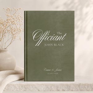 Può includere: Un libro verde scuro con le parole "The Officiant" e "John Black" in corsivo bianco. I nomi "Emma & James" e la data "25.06.2026" sono stampati sulla copertina. Il libro è su una superficie chiara.