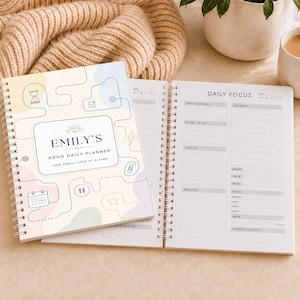 Puede incluir: Una agenda diaria de espiral con una cubierta de tonos pastel que presenta formas abstractas, iconos de líneas y el texto 'EMILY'S ADHD DAILY PLANNER ONE SMALL STEP AT A TIME'. La página abierta muestra un diseño 'DAILY FOCUS' con secciones.