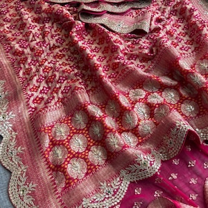 May include: A vibrant fuchsia and red saree featuring intricate gold zari embroidery, a repeating geometric pattern, and circular floral motifs. The fabric is adorned with a scalloped border embellished with gold thread, sequins, and small green accents.