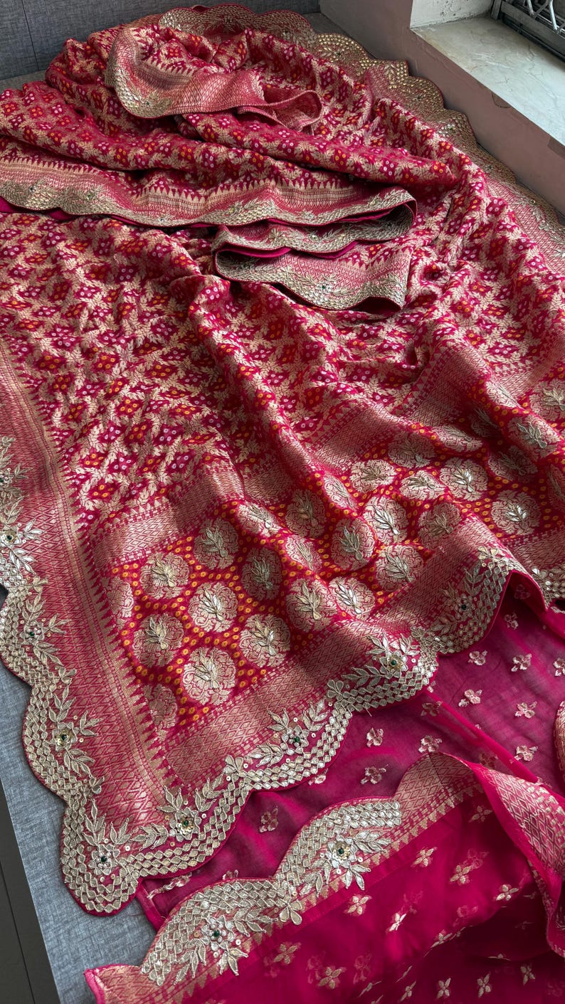 May include: A vibrant fuchsia and red saree featuring intricate gold zari embroidery, a repeating geometric pattern, and circular floral motifs. The fabric is adorned with a scalloped border embellished with gold thread, sequins, and small green accents.