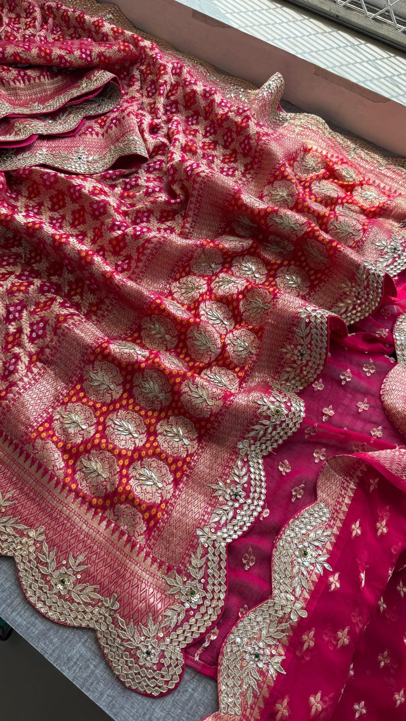 May include: A vibrant fuchsia and red textile featuring intricate gold metallic embroidery in circular floral patterns. The fabric displays a traditional tie-dye effect with small yellow dots and a scalloped border embellished with gold thread.