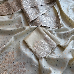 May include: A cream-coloured fabric featuring a scattered pattern of small dark and orange dots. The material is adorned with wide, scalloped borders decorated with intricate gold-toned floral embroidery and metallic sequins throughout the design.