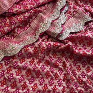May include: A vibrant magenta fabric featuring a repeating gold floral pattern and small white and yellow dot accents. The textile has a scalloped edge border embellished with intricate gold embroidery, metallic sequins, and small green stones.