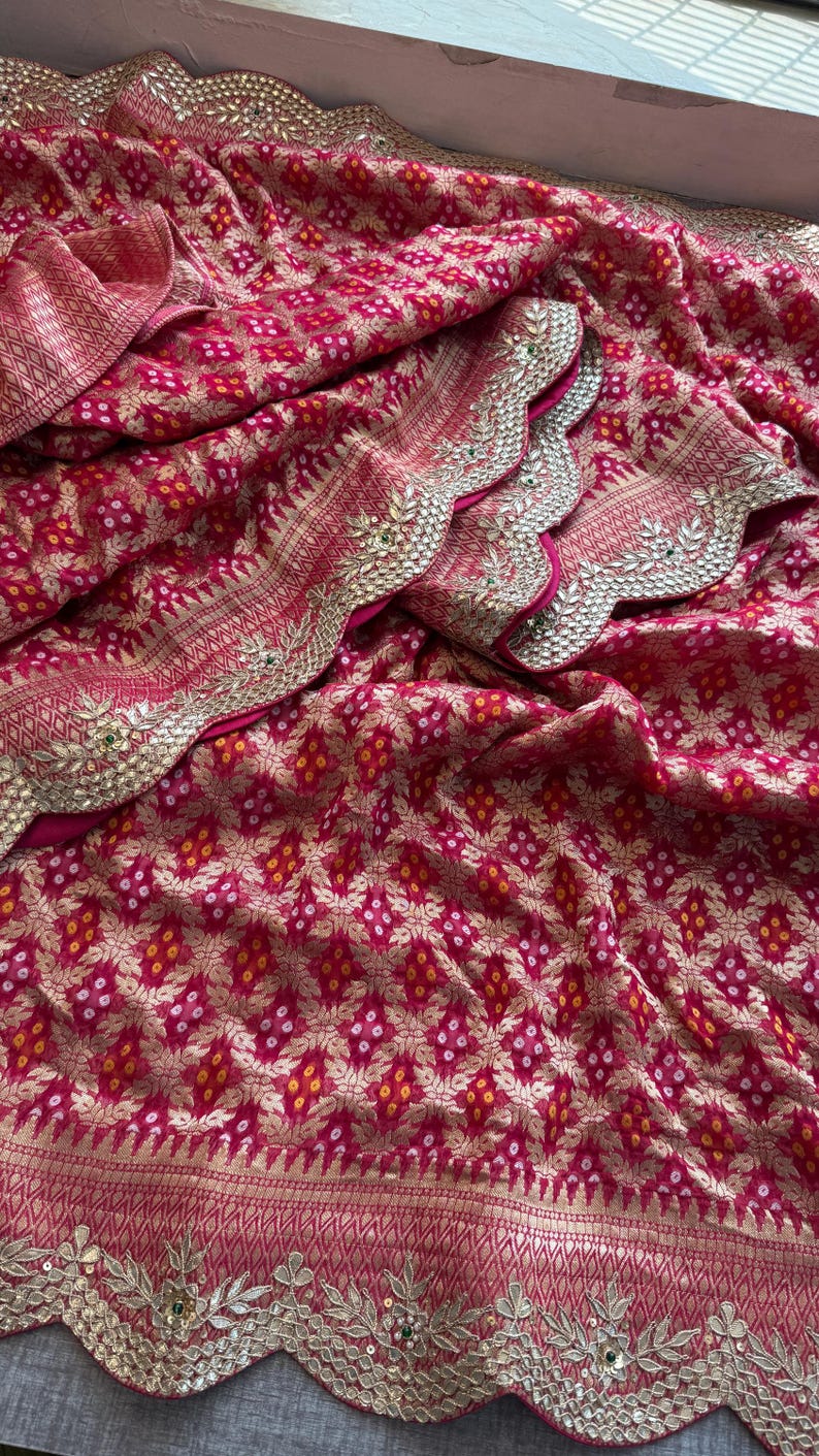 May include: A vibrant magenta fabric featuring a repeating gold floral pattern and small white and yellow dot accents. The textile has a scalloped edge border embellished with intricate gold embroidery, metallic sequins, and small green stones.