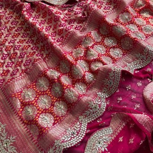 May include: A vibrant fuchsia and red saree featuring a dense gold zari woven pattern with circular floral motifs. The fabric is adorned with a scalloped border embellished with intricate silver metallic embroidery and small reflective mirror accents.