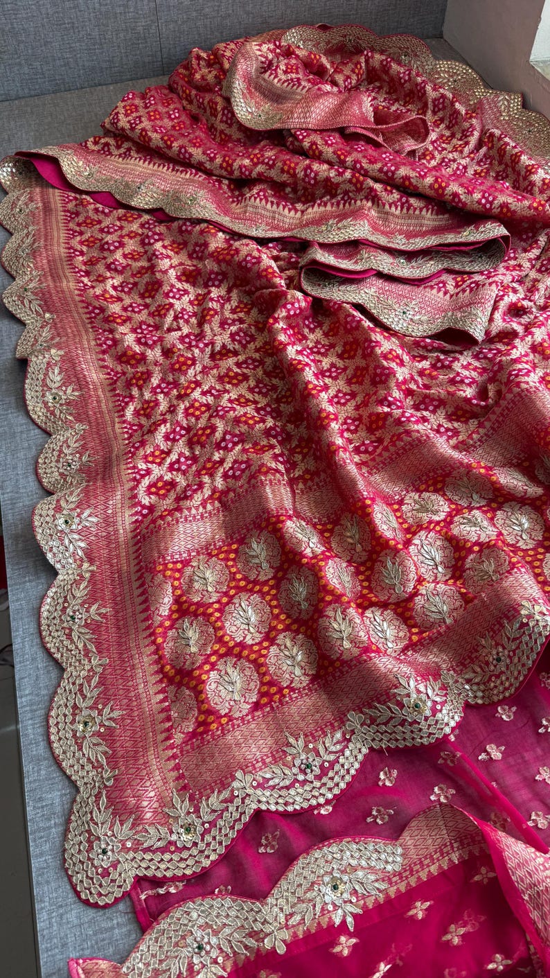 May include: A vibrant fuchsia and red fabric featuring a dense, intricate pattern of small white dots and gold metallic floral motifs. The textile is finished with a wide, scalloped border embellished with detailed gold embroidery and sequins.