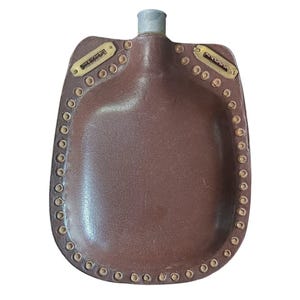 Puede incluir: Una cantimplora cubierta de cuero marrón con una boquilla de metal plateado. La cantimplora presenta un borde cosido con una serie de pequeños ojales metálicos circulares a lo largo del borde y dos placas metálicas rectangulares cerca del cuello.