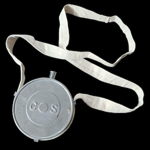 May include: A circular silver-toned metal canteen featuring the embossed text COS on the front face. The canteen includes a small metallic spout at the top and is attached to a long, off-white fabric shoulder strap made of woven material.