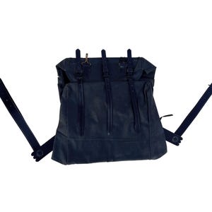 May include: A dark navy blue canvas backpack featuring three vertical straps with buckles on the front flap. The bag includes two adjustable shoulder straps attached to the sides, each with a metal triangular ring at the top end for secure closure.