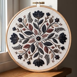 May include: A circular wooden embroidery hoop containing a piece of off-white fabric with a hand-stitched floral mandala design. The embroidery features dark navy, rust-colored, and beige thread depicting symmetrical flowers, leaves, and ferns.