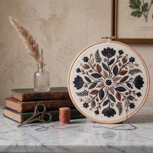May include: A circular wooden embroidery hoop containing a cream-colored fabric with a symmetrical floral and leaf pattern in shades of dark blue, brown, and tan. A needle with thread is attached to the fabric, alongside vintage scissors.