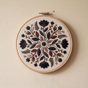 May include: A circular wooden embroidery hoop frames a piece of light beige fabric featuring a symmetrical floral design. The embroidery consists of dark navy, rust orange, and tan thread depicting various leaves, ferns, and flowers.