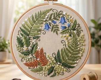 May include: A circular wooden embroidery hoop containing a beige fabric panel with a hand-stitched wreath design. The wreath features green and rust-colored fern fronds, blue bell-shaped flowers, and small yellow blossoms with white accents.