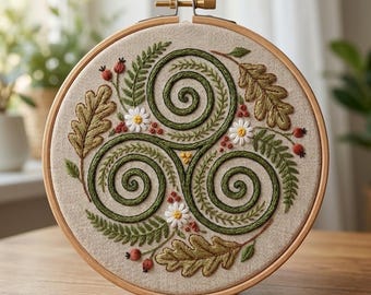 May include: A circular wooden embroidery hoop containing a beige fabric panel with a hand-stitched triskelion design. The pattern features dark green embroidered spirals surrounded by oak leaves, fern fronds, small white flowers, and red berries.