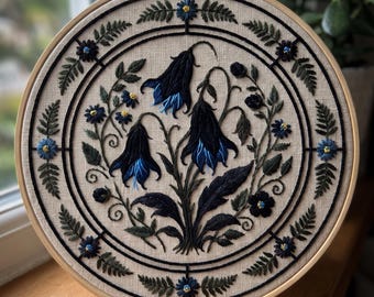 May include: A circular embroidery hoop containing a beige fabric panel with a hand-stitched design of three large, dark blue bell-shaped flowers surrounded by dark green fern leaves and smaller blue blossoms, all framed by a black circular border.