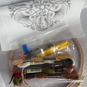 May include: An embroidery kit featuring a white fabric panel with a black floral line art design, a wooden hoop, a yellow seam ripper, a small pink flower-shaped needle minder, and a collection of embroidery floss in brown, green, red, and white.
