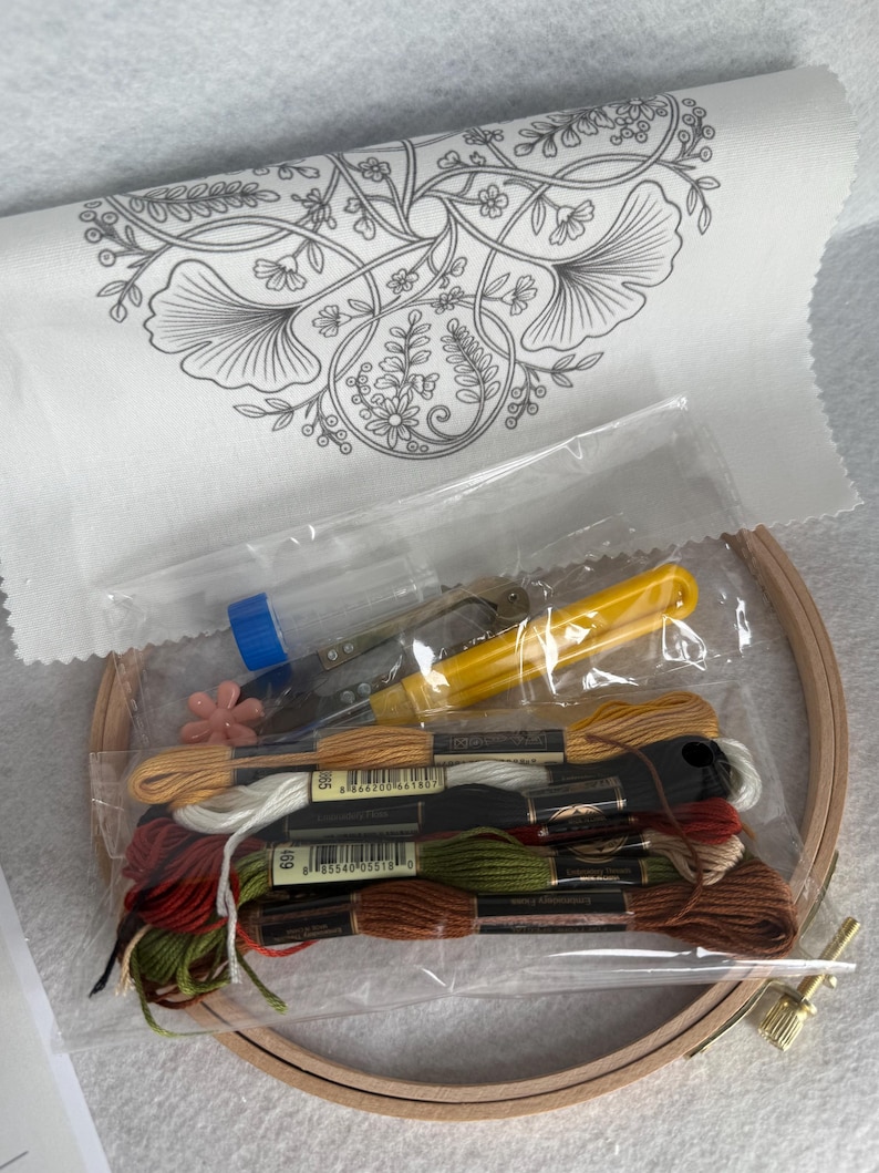 May include: An embroidery kit featuring a white fabric panel with a black floral line art design, a wooden hoop, a yellow seam ripper, a small pink flower-shaped needle minder, and a collection of embroidery floss in brown, green, red, and white.