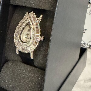 May include: A teardrop-shaped wristwatch featuring a silver-tone case encrusted with baguette-cut and round clear stones surrounding a white dial. The watch has a black strap and is displayed within a black foam-lined presentation box.