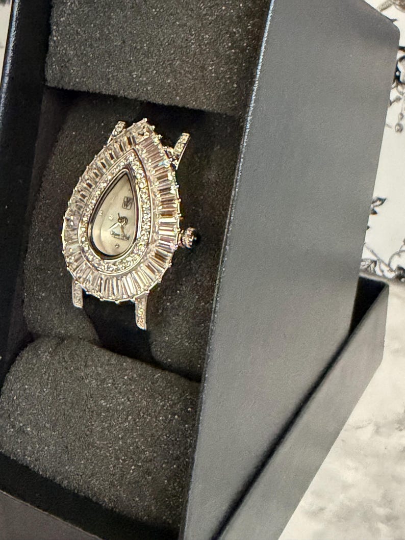 May include: A teardrop-shaped wristwatch featuring a silver-tone case encrusted with baguette-cut and round clear stones surrounding a white dial. The watch has a black strap and is displayed within a black foam-lined presentation box.