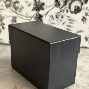 May include: A rectangular black box with a lid, featuring a smooth, matte finish. The box is structured with sharp, clean edges and a solid, opaque black color throughout its exterior surface, presenting a simple and functional design.