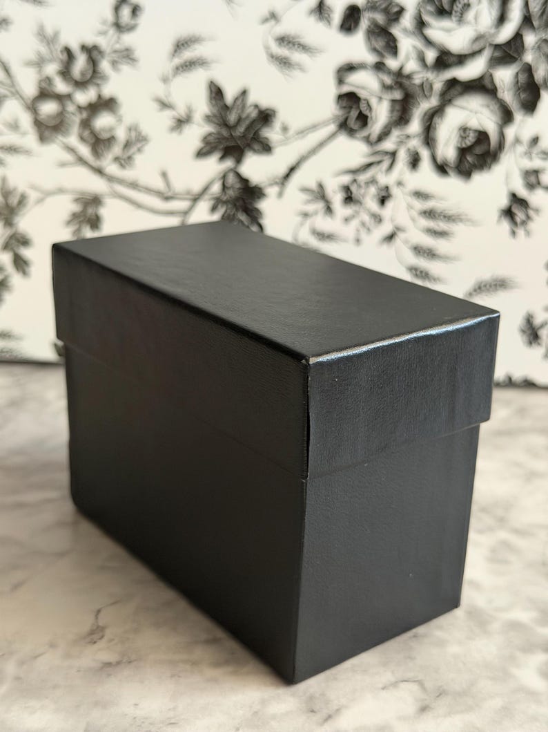 May include: A rectangular black box with a lid, featuring a smooth, matte finish. The box is structured with sharp, clean edges and a solid, opaque black color throughout its exterior surface, presenting a simple and functional design.
