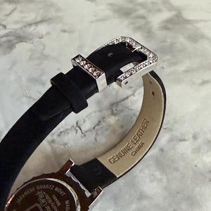 May include: A black watch strap with a silver-tone buckle encrusted with clear crystals. The inner side of the strap is beige and stamped with the text Genuine Leather and China. The watch case back is silver-tone with text including Japanese Quartz Movt.