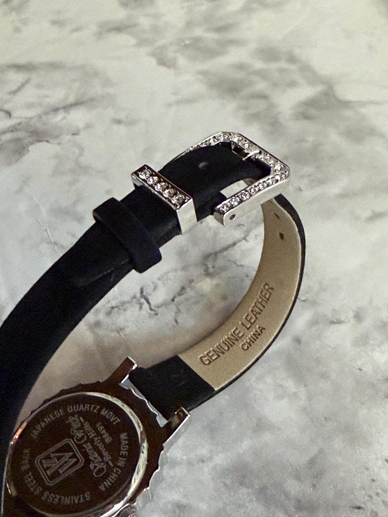 May include: A black watch strap with a silver-tone buckle encrusted with clear crystals. The inner side of the strap is beige and stamped with the text Genuine Leather and China. The watch case back is silver-tone with text including Japanese Quartz Movt.