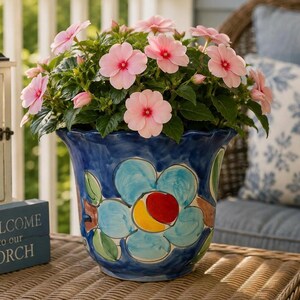 May include: A ceramic planter with a scalloped rim, featuring a hand-painted blue floral design with a central red and yellow accent. The planter is filled with a lush plant displaying numerous light pink flowers with dark pink centers.