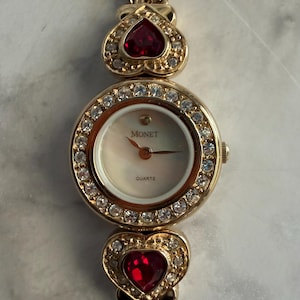 May include: A gold-toned Monet quartz watch with a round face and a bezel encrusted with clear crystals. The watch features a cream-colored dial and is accented with red heart-shaped jewels. The watch band is also gold-toned.