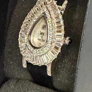 May include: A teardrop-shaped wristwatch with a silver-tone case and a white dial. The bezel is encrusted with numerous baguette-cut and round-cut clear stones. The watch features a crown on the right side and a black strap, presented in a dark case.