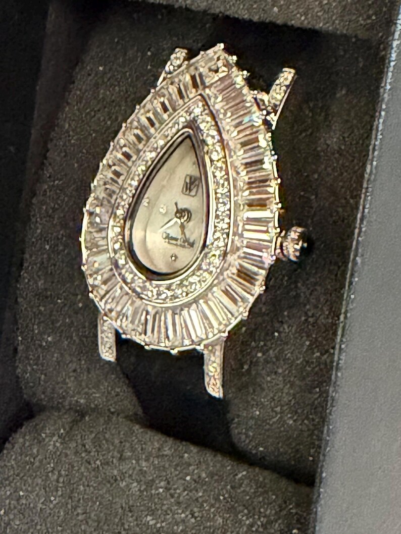 May include: A teardrop-shaped wristwatch with a silver-tone case and a white dial. The bezel is encrusted with numerous baguette-cut and round-cut clear stones. The watch features a crown on the right side and a black strap, presented in a dark case.