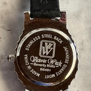 May include: The back of a silver-tone watch with a black leather strap. The case back is engraved with the text: STAINLESS STEEL BACK, a logo with the letters VW, Victoria Wieck Beverly Hills, B6491, MADE IN CHINA, and JAPANESE QUARTZ MOVT.