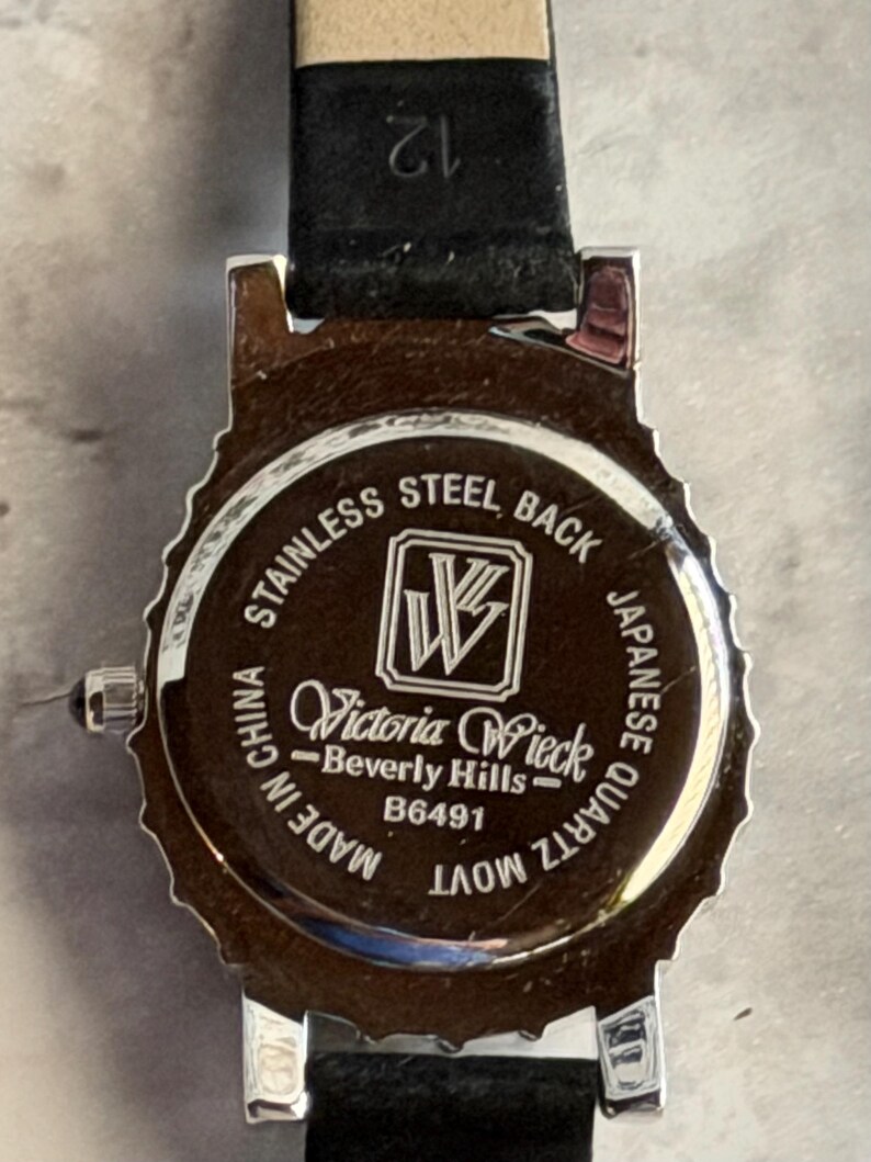 May include: The back of a silver-tone watch with a black leather strap. The case back is engraved with the text: STAINLESS STEEL BACK, a logo with the letters VW, Victoria Wieck Beverly Hills, B6491, MADE IN CHINA, and JAPANESE QUARTZ MOVT.