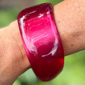 May include: A wide, translucent fuchsia-colored bangle bracelet with a smooth, glossy finish. The bracelet features a rounded, chunky profile and a vibrant, deep pink hue that catches the light, creating subtle reflections across its curved surface.