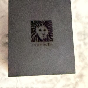 May include: A rectangular dark gray box featuring a centered square logo with a stylized lion head graphic and the text Anne Klein printed in black. The box has a matte finish and a clean, simple design with sharp edges and a flat top surface.