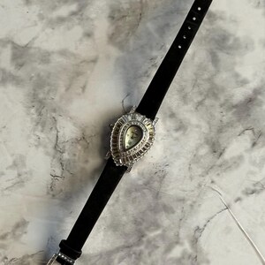 May include: A wristwatch with a black strap and a teardrop-shaped silver-tone case. The bezel is encrusted with baguette-cut clear crystals surrounding a light-colored dial. The buckle on the black strap is also decorated with clear crystals.