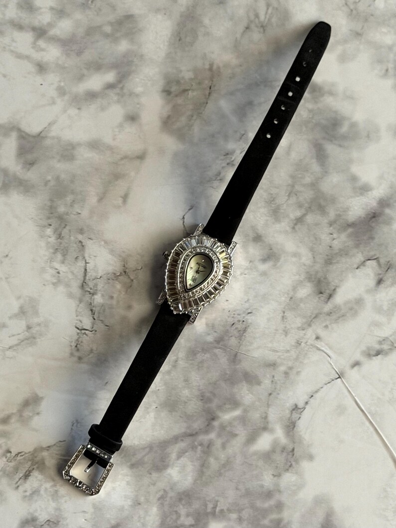 May include: A wristwatch with a black strap and a teardrop-shaped silver-tone case. The bezel is encrusted with baguette-cut clear crystals surrounding a light-colored dial. The buckle on the black strap is also decorated with clear crystals.