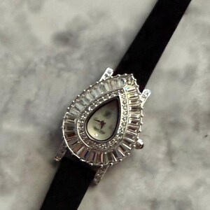 May include: A silver-toned wristwatch featuring a teardrop-shaped case surrounded by a border of baguette-cut clear crystals. The watch face is white with thin hands and is attached to a slim, solid black strap that extends vertically.